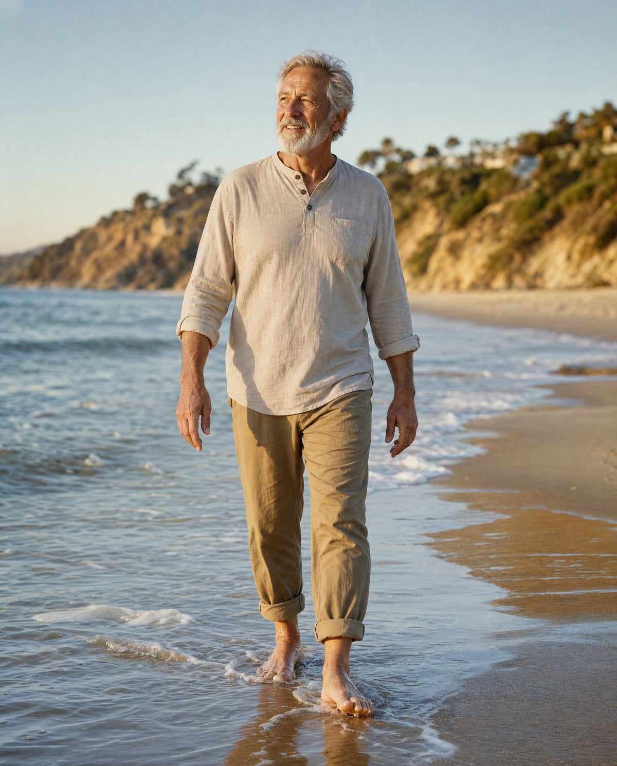 Man in his seventies walking barefoot along the edge of the water on a sandy beach at golden hour